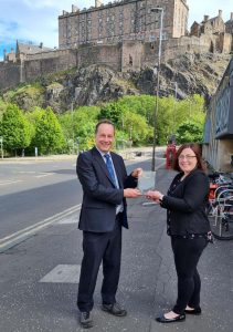 Fiona from SUSE presenting the Inclusive Workplace Award to the Vice-Principal of The University of Edinburgh. Standing in a quiet street in front of Edinburgh Castle.