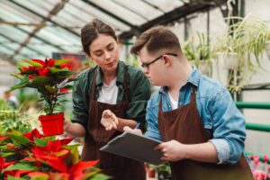 Two people working in a garden centre. One is holding a red potted plant and the other is holding a clipboard. The first person is talking and gesturing to the clipboard.