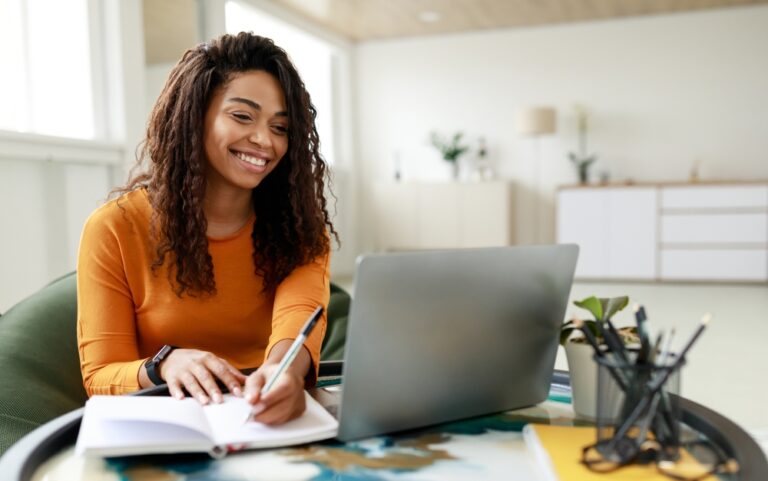 A photo of a person sitting at a desk, smiling at a laptop screen and holding a pen ready to write in a notebook.