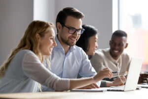 Two people working together at a laptop and smiling. There are another two people in the background.