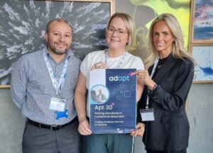 Three members of NHS 24 staff standing, smiling at the camera, holding an Apt 30 poster.