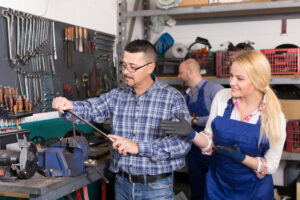Two people in a workshop. One person is using equipment and the other is looking on and helping.